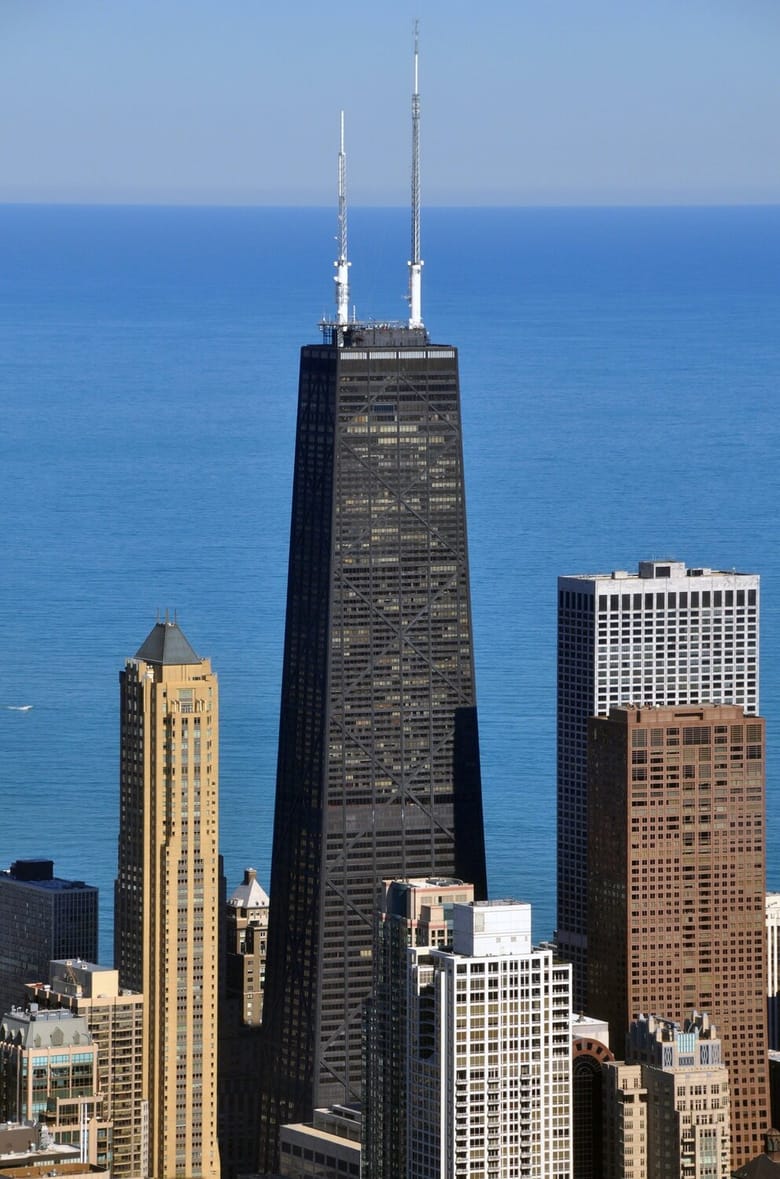 Aerial shot of 875. North Michigan Avenue, a black mid-century modern building with two tall white antennas, surrounded by other builings